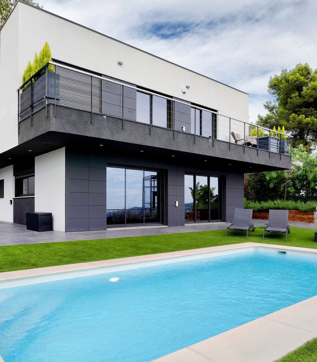 Moderna casa blanca y gris con piscina, balcón y césped bajo un cielo nublado.