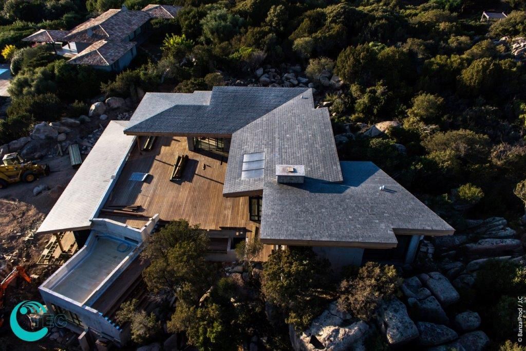 Vue aérienne d'une maison moderne avec toit gris, terrasse en bois et arbres environnants.