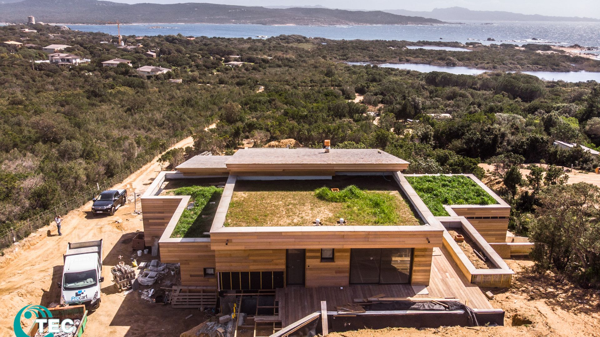 Maison moderne en bois avec toit végétalisé, vue sur l'océan, avec des engins de chantier à proximité.