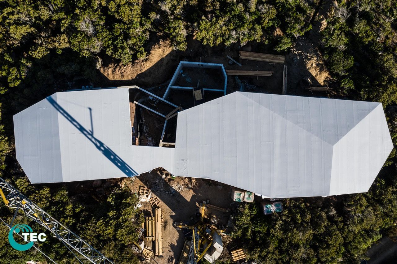 Vue aérienne d'une maison moderne en construction, au toit blanc, sur un flanc de colline boisé.