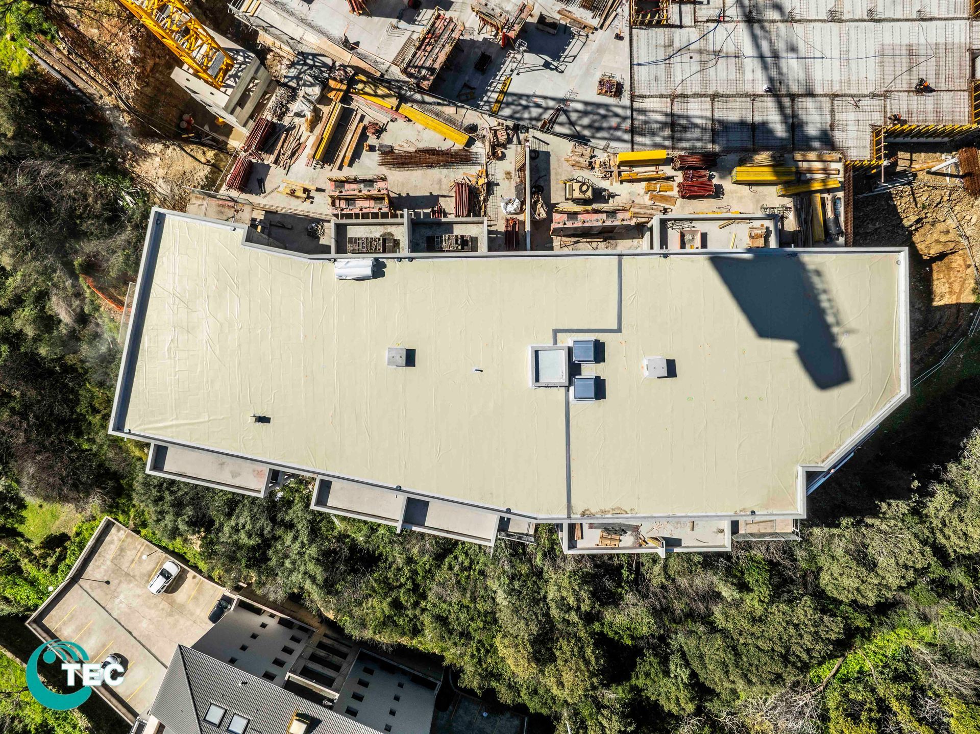 Vue aérienne du toit d'un bâtiment entouré de matériaux de construction ; à proximité d'arbres et d'un autre bâtiment.