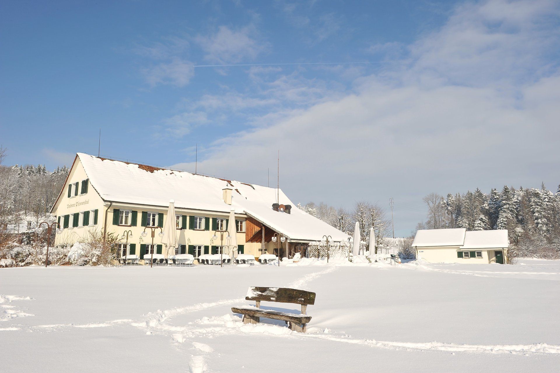 Ein schneebedecktes Feld mit einem Gebäude im Hintergrund und einer Bank im Vordergrund. Foto von der Restaurant Pfannenstiel AG.