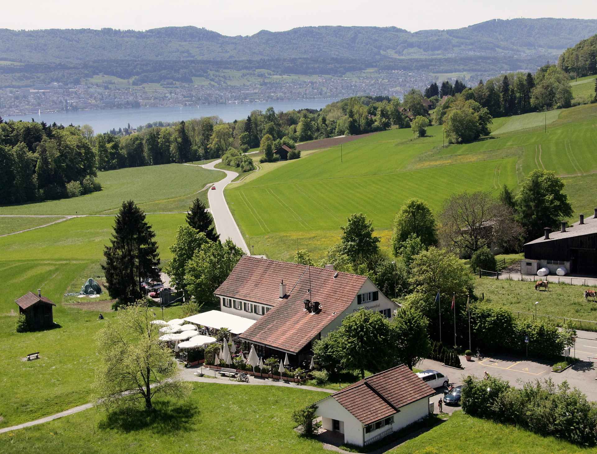 Luftaufnahme eines Gebäudes inmitten eines Feldes mit einem See im Hintergrund. Foto von der Restaurant Pfannenstiel AG.