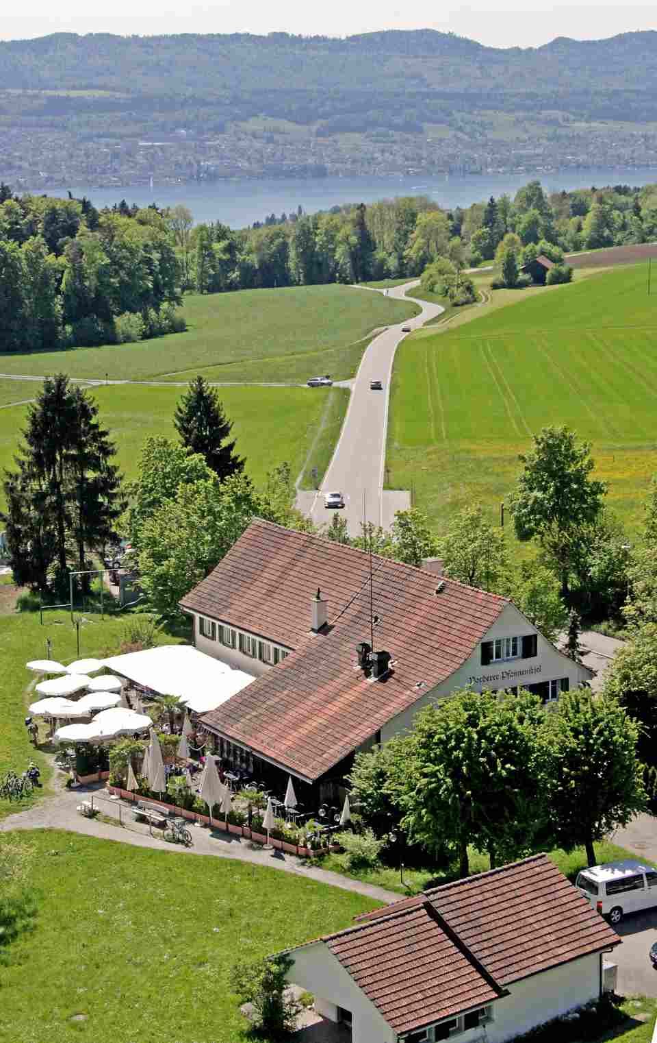 Luftaufnahme eines Gebäudes inmitten eines Feldes mit einem See im Hintergrund. Foto von der Restaurant Pfannenstiel AG.