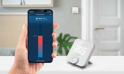 Eine Person hält ein Smartphone neben einem Thermostat.