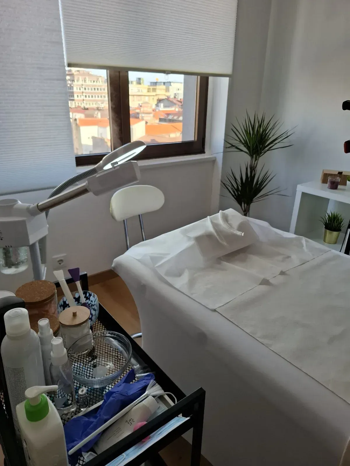 Sala de tratamientos de spa luminosa con camilla de masaje, lámpara, carrito de suministros y ventana grande.