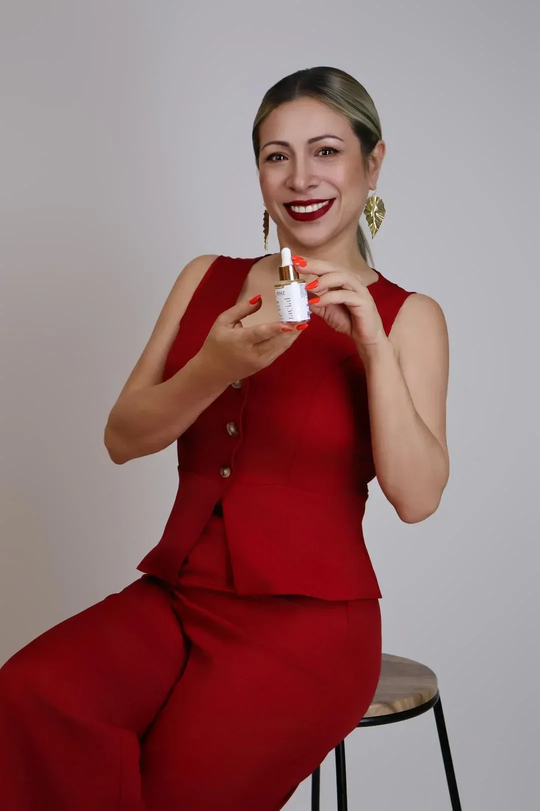 Mujer con atuendo rojo sosteniendo una pequeña botella de producto y sonriendo mientras está sentada