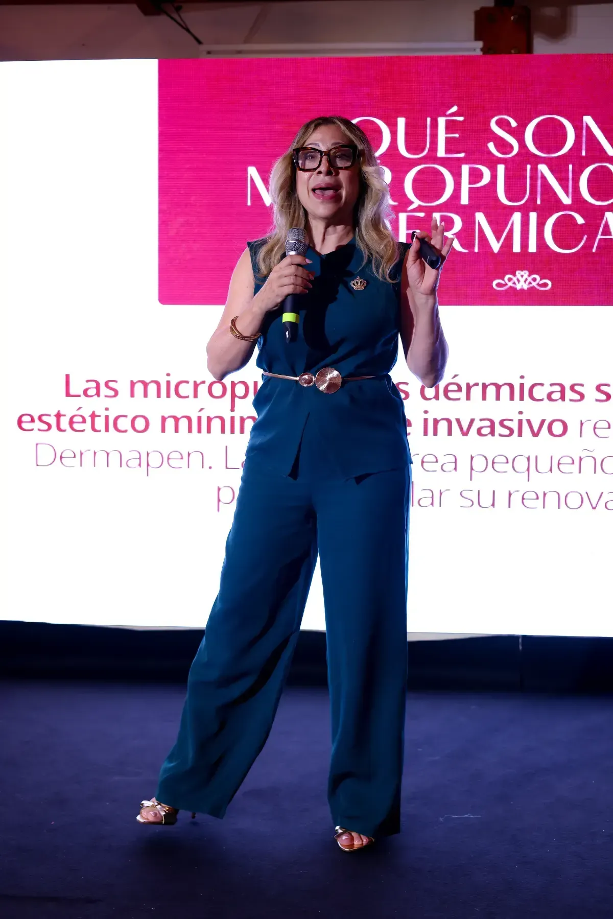 Mujer hablando en el escenario con micrófono frente a una pantalla de presentación rosa.