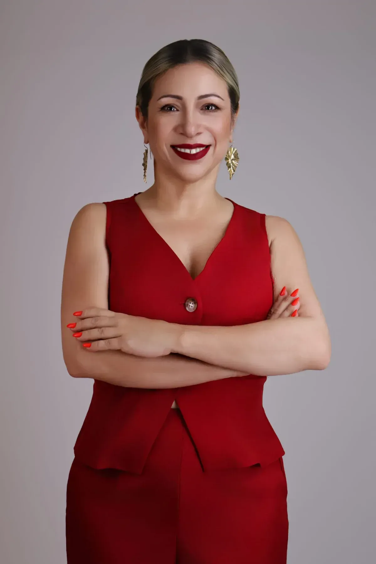 Mujer con traje rojo de pie con los brazos cruzados y sonriendo sobre un fondo gris.