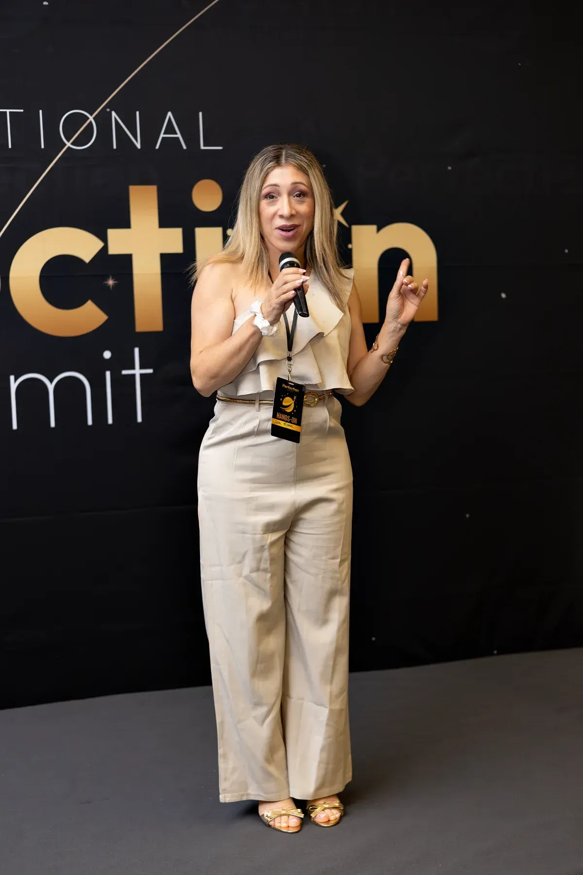 Mujer hablando en un escenario con atuendo beige, sosteniendo un micrófono frente a un fondo de conferencia.