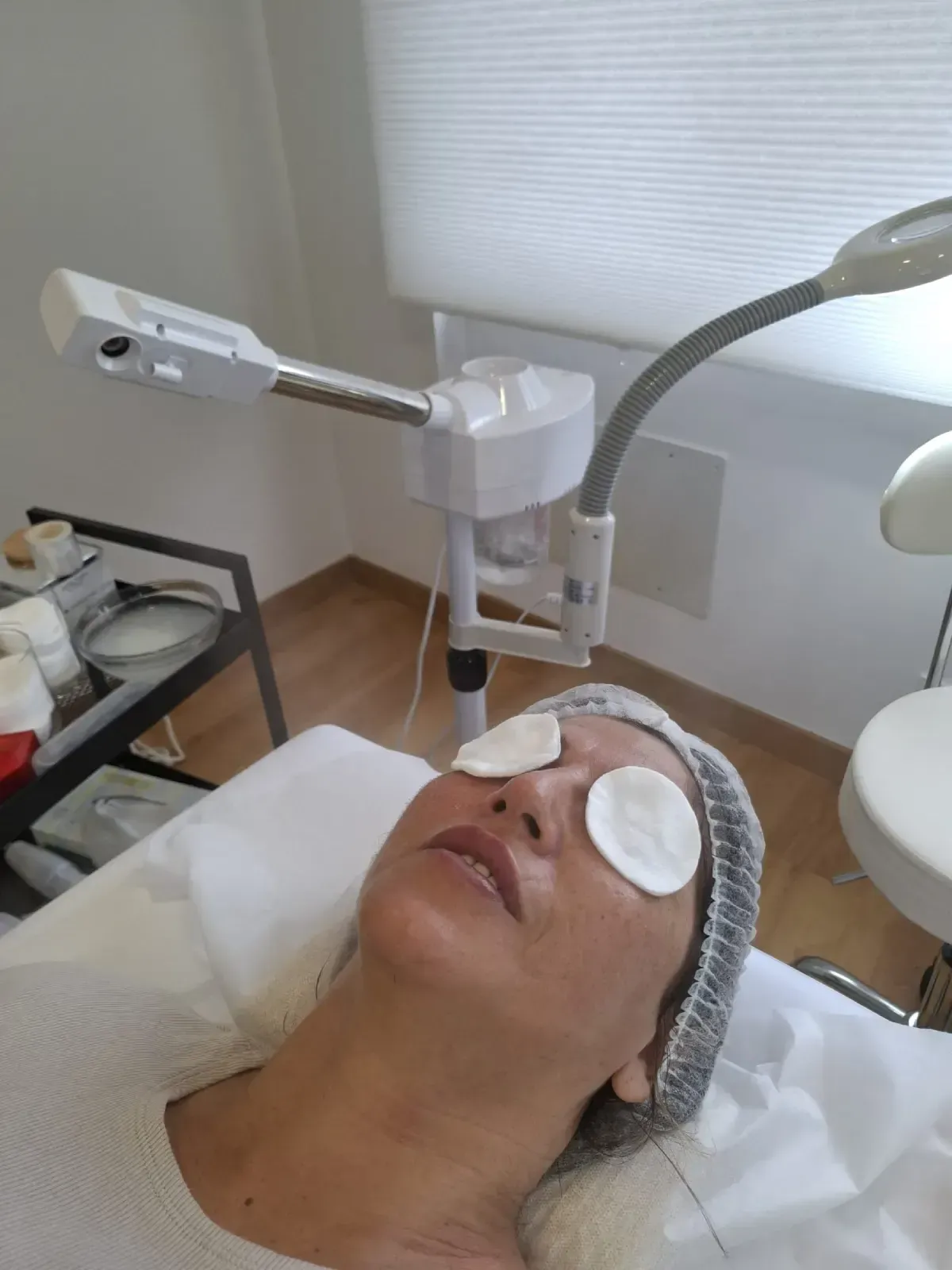 Persona recibiendo un tratamiento facial en una sala de spa, con parches para los ojos y un dispositivo de tratamiento cerca.