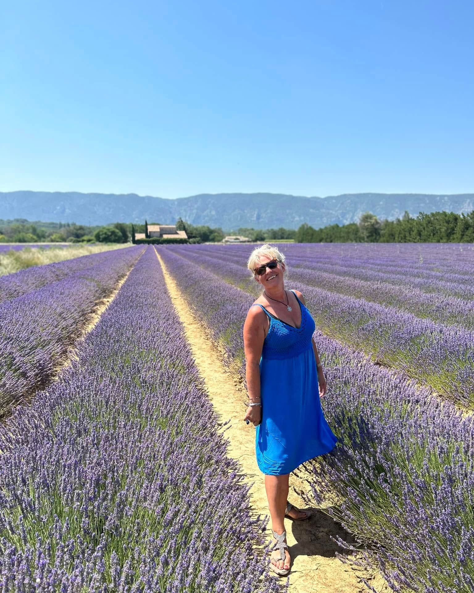 Flore en robe bleue debout dans un champ de lavande, avec des montagnes et un ciel bleu en arrière-plan.