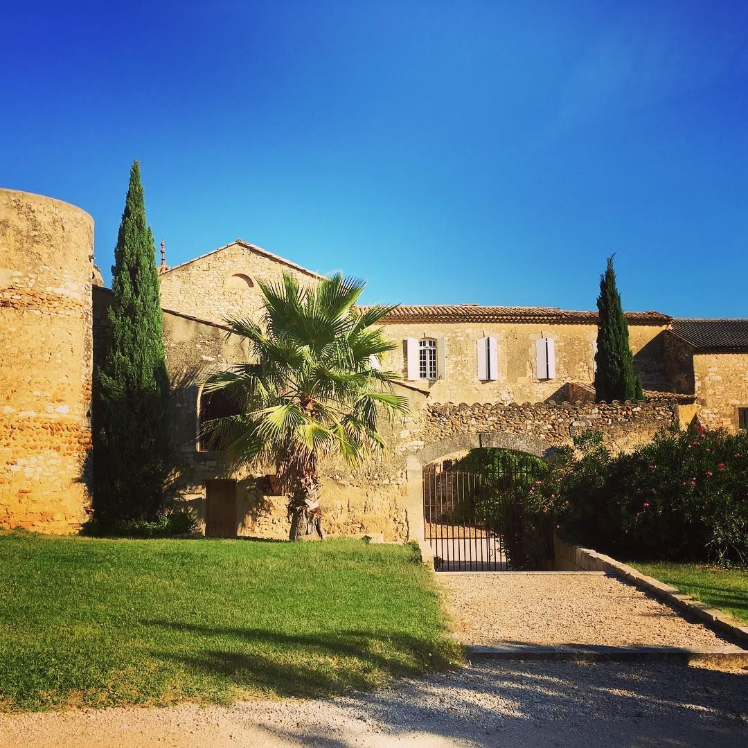 Cour en pierre et bâtiment rustique avec portail arqué, cyprès et palmiers sous un ciel d'un bleu éclatant