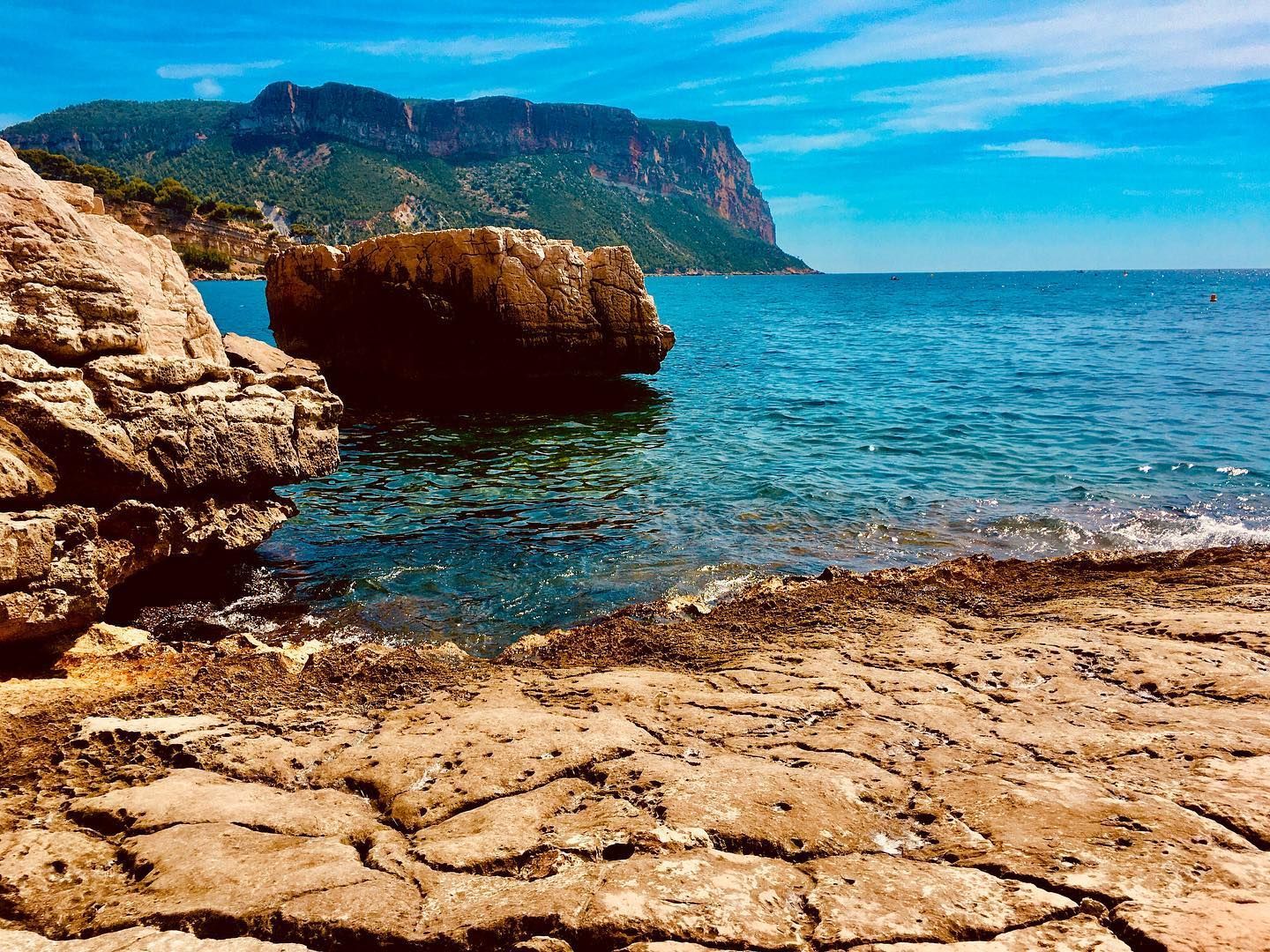 Falaise rocheuse en bord de mer avec eau turquoise, ciel bleu et montagnes au loin sous un soleil éclatant
