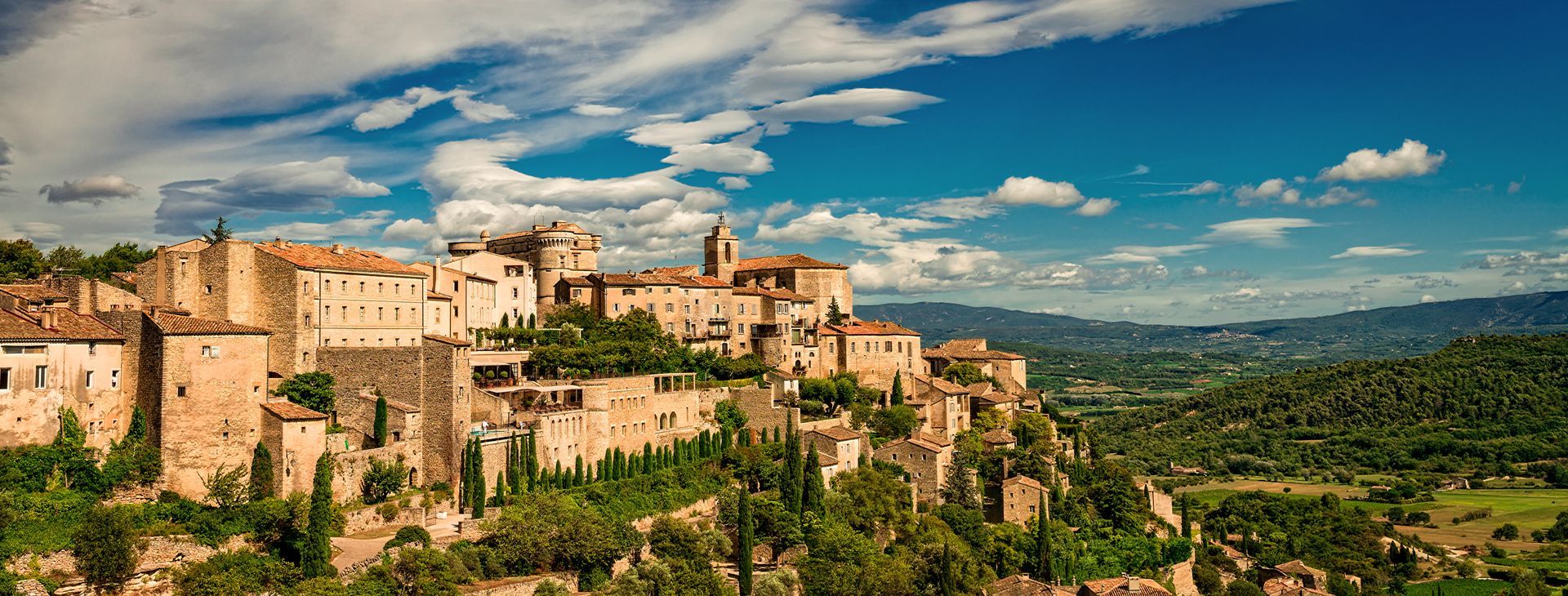 Village, toits de tuiles et collines verdoyantes sous un ciel bleu parsemé de nuages