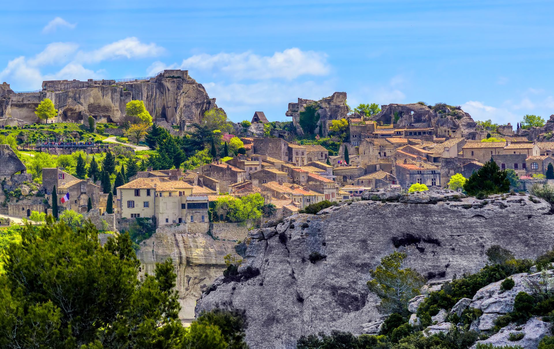 Ruines de pierre antiques sur une colline sous un ciel bleu, entourées d'arbres verdoyants et de falaises rocheuses.