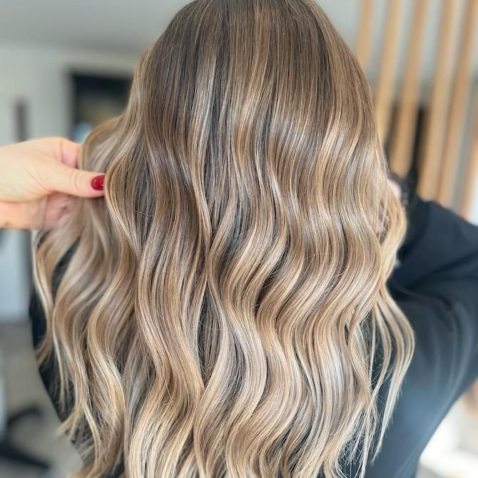 Cheveux blonds et châtain clair ondulés avec des mèches, coiffés en boucles souples. Une main caresse les cheveux.