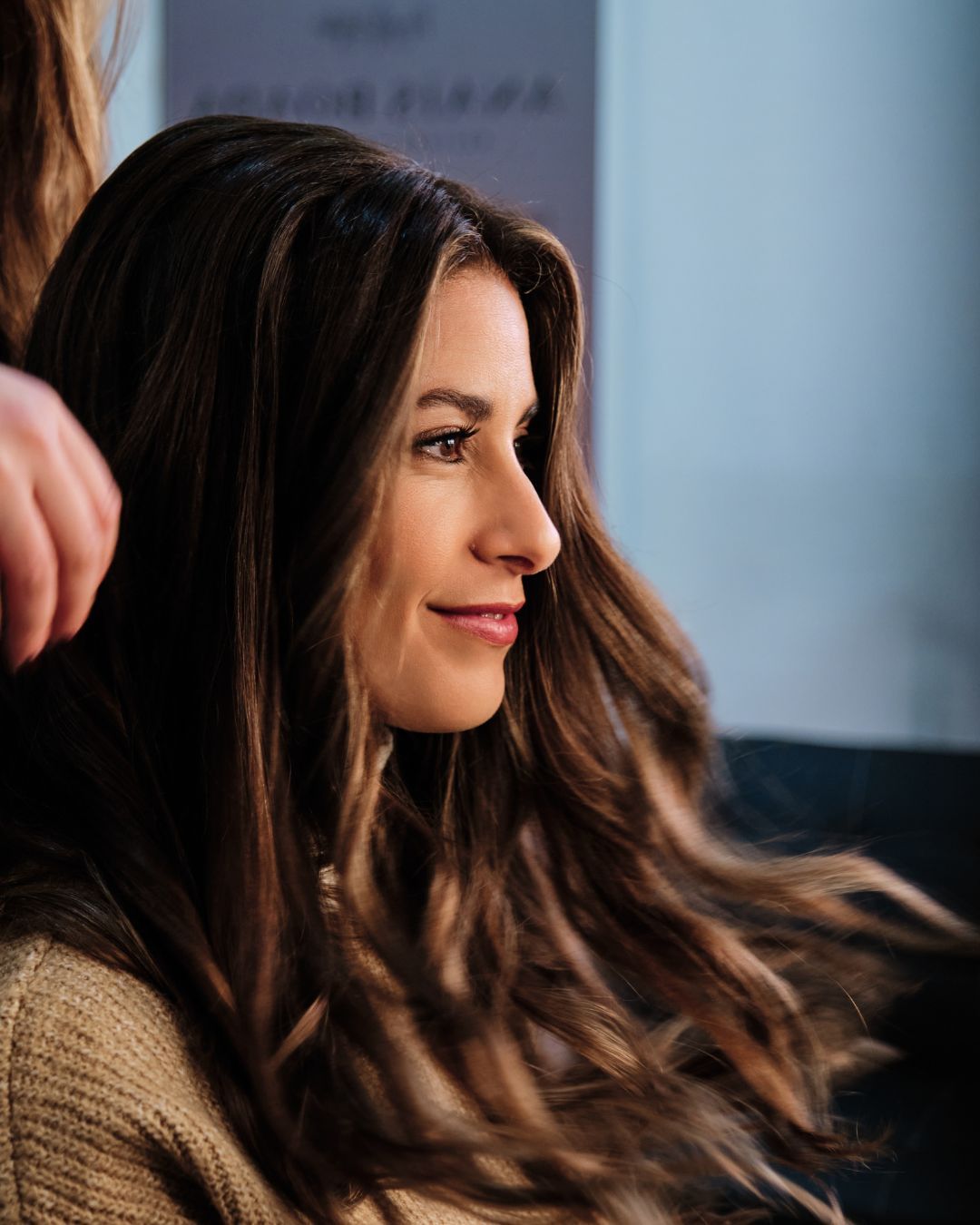 Femme aux longs cheveux bruns ondulés, regardant sur le côté, dans un salon de coiffure ; en train de se faire coiffer.