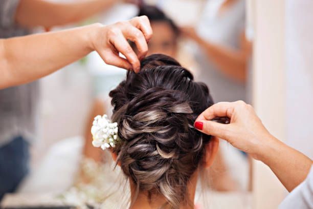 Des mains coiffent une femme aux cheveux foncés relevés, ornés d'une fleur blanche, devant un miroir.