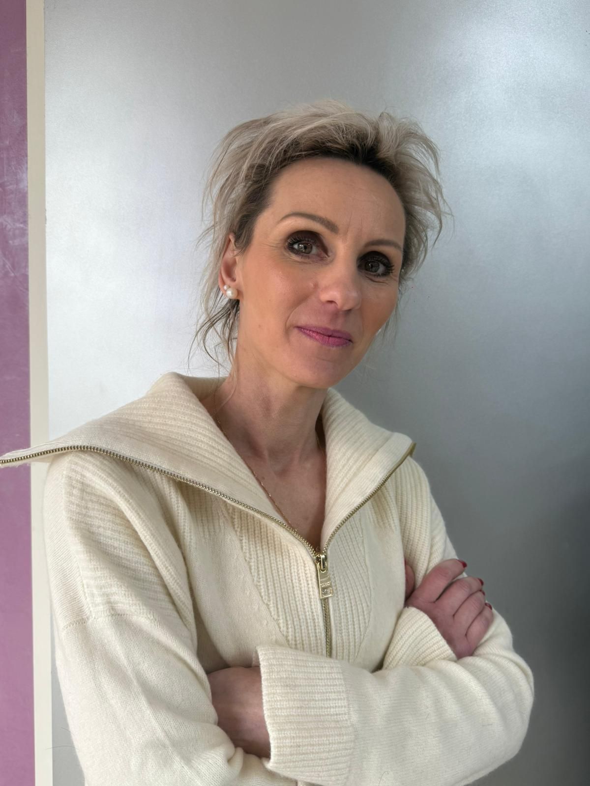 Femme dans un salon de coiffure, les bras croisés, souriante. Elle porte un pull bordeaux, un jean, une ceinture noire et a les cheveux blonds courts.