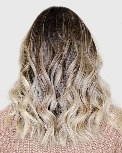 Cheveux blonds ondulés balayés avec des racines foncées, vus de dos sur un fond clair.