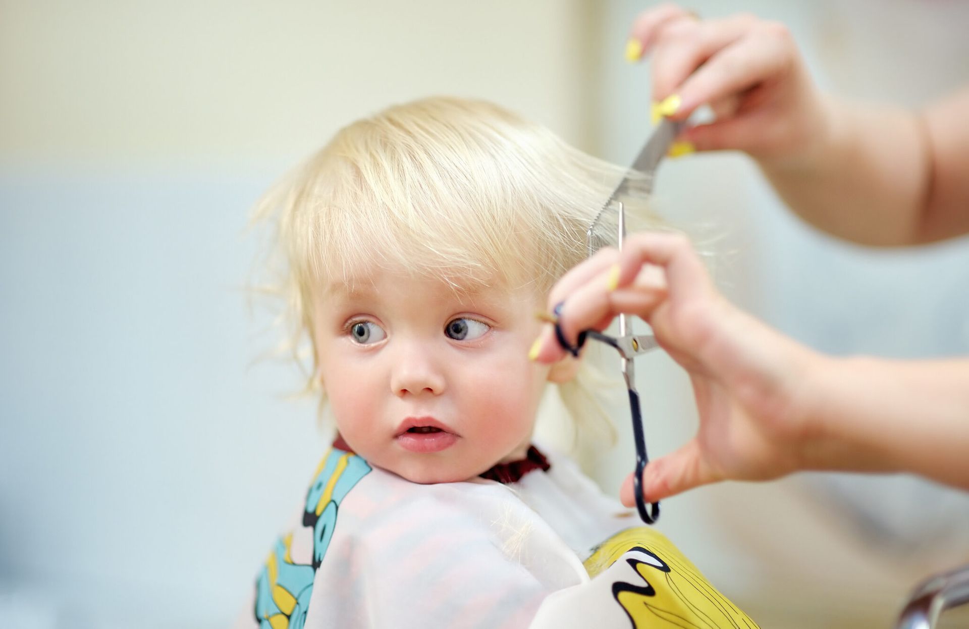 Petite fille blonde aux grands yeux se fait couper les cheveux ; ses mains tiennent des ciseaux.