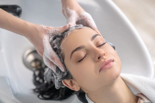 Une femme se fait laver les cheveux, les yeux fermés, dans un salon de coiffure. Des mains massent ses cheveux savonneux. Fond blanc.