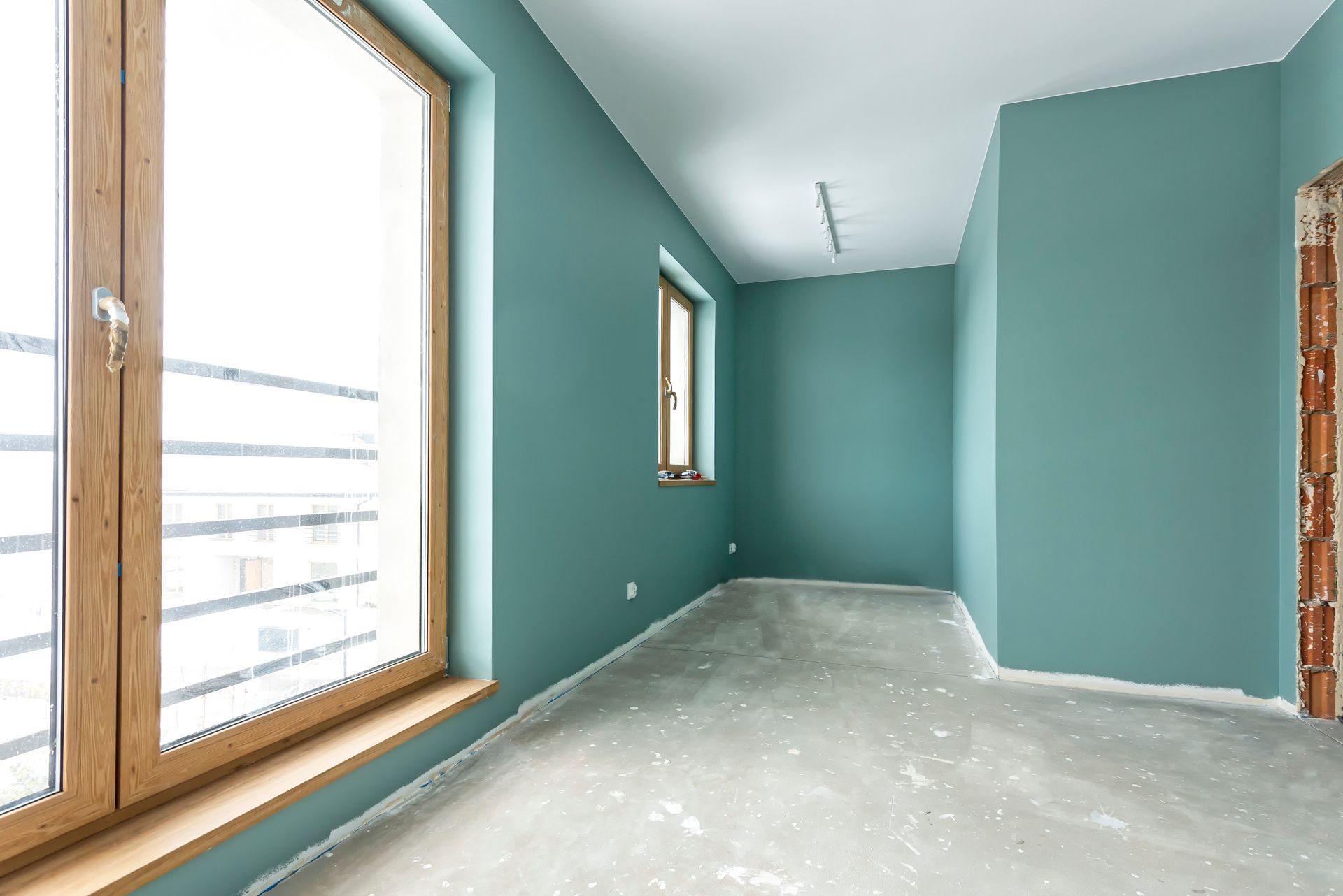 Une pièce intérieure en construction, avec des murs turquoise, une grande fenêtre et un sol en béton brut.
