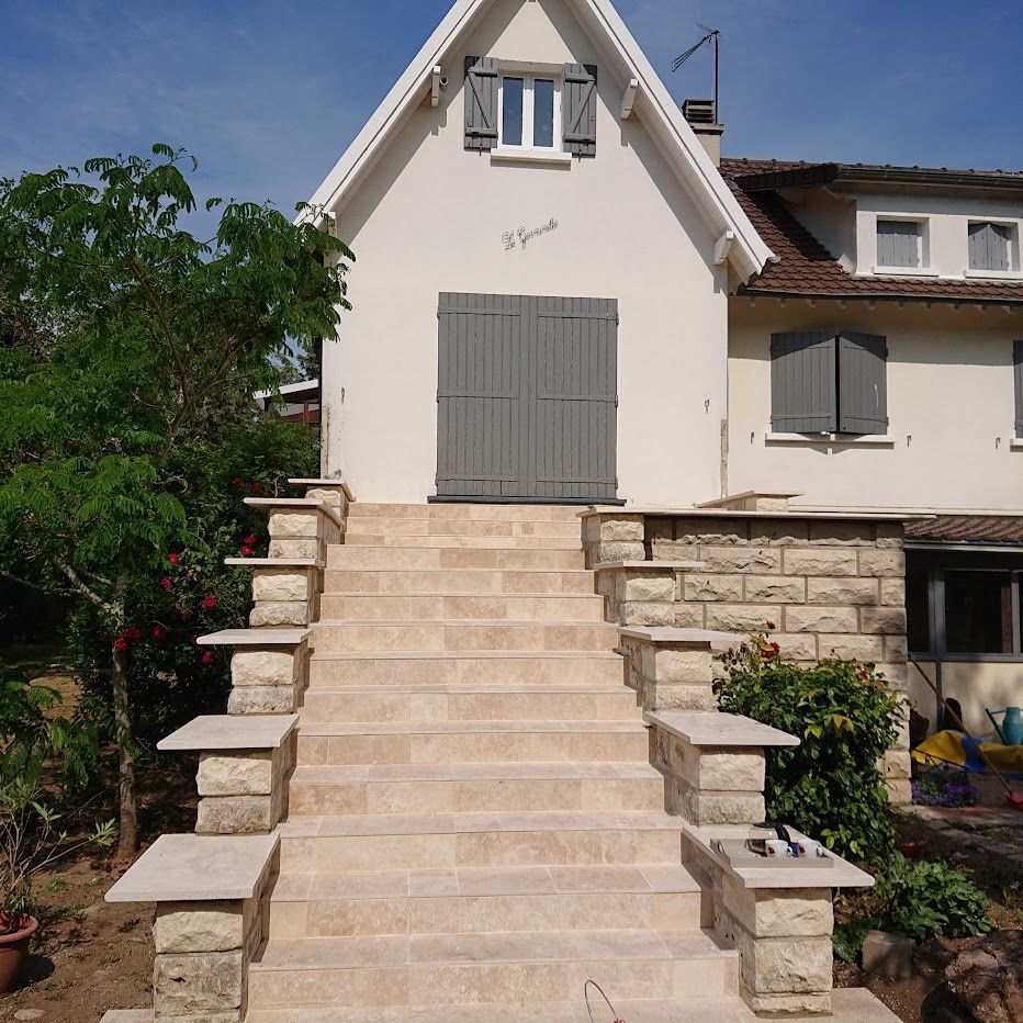 Un large escalier en pierre mène à une maison aux tons clairs, avec une porte en bois et des volets gris.