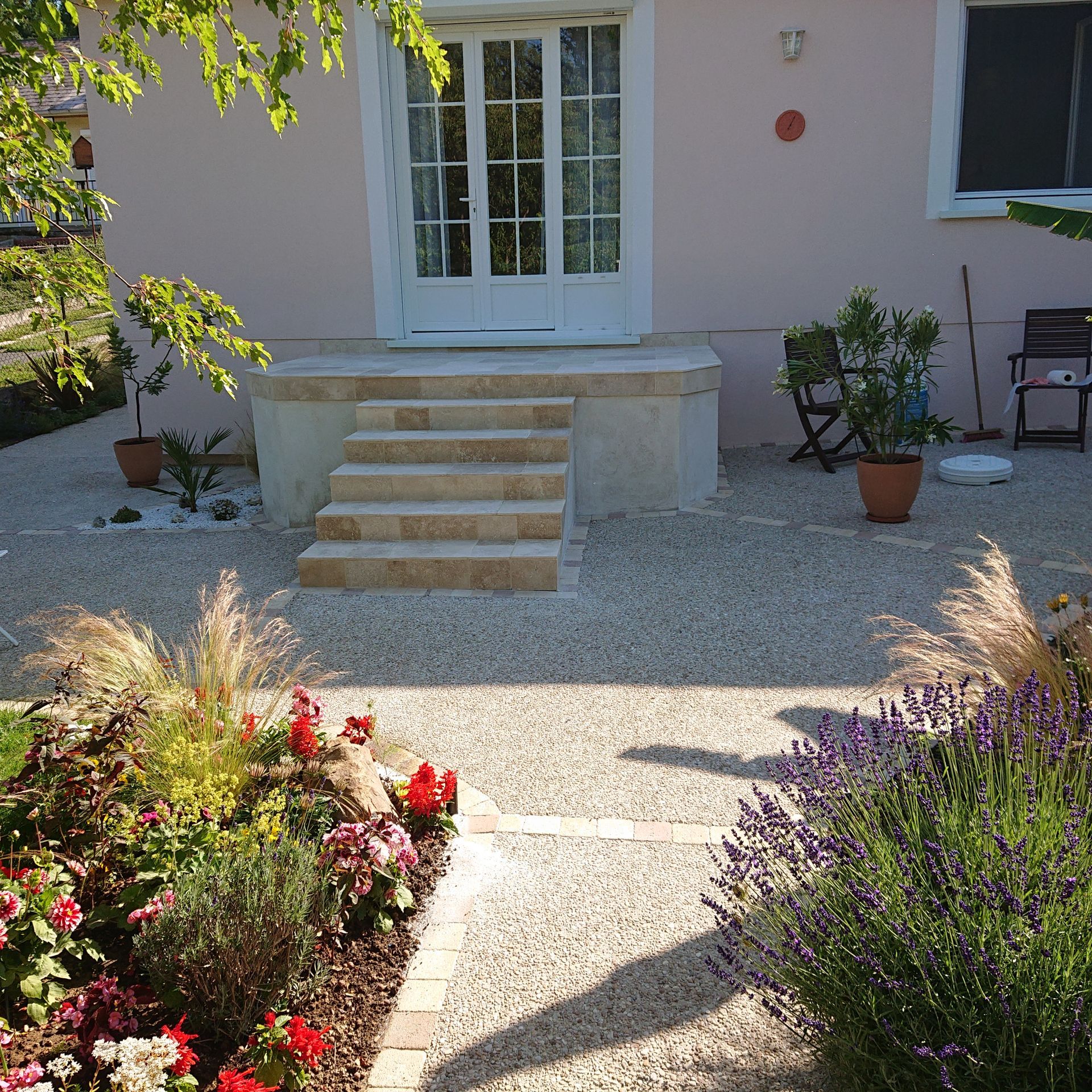 Un escalier en pierre mène à des portes-fenêtres blanches donnant sur une maison rose pâle, entourée de gravier.