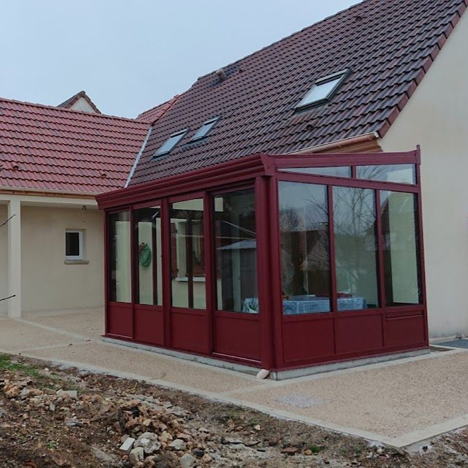 Une véranda de couleur brun rougeâtre, adossée à une maison de couleur claire.