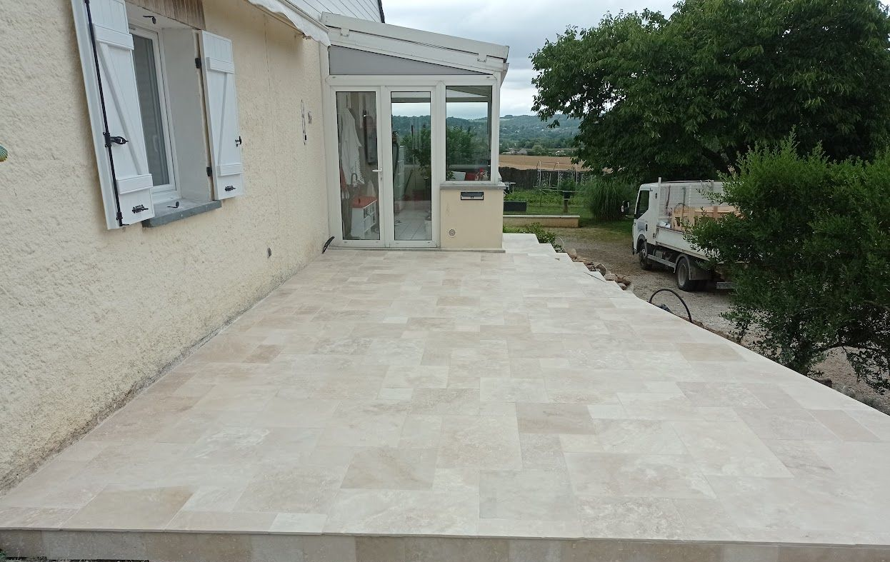 Couverture de toit Patio carrelé beige à l'extérieur d'un bâtiment de couleur claire avec portes vitrées et volets blancs.