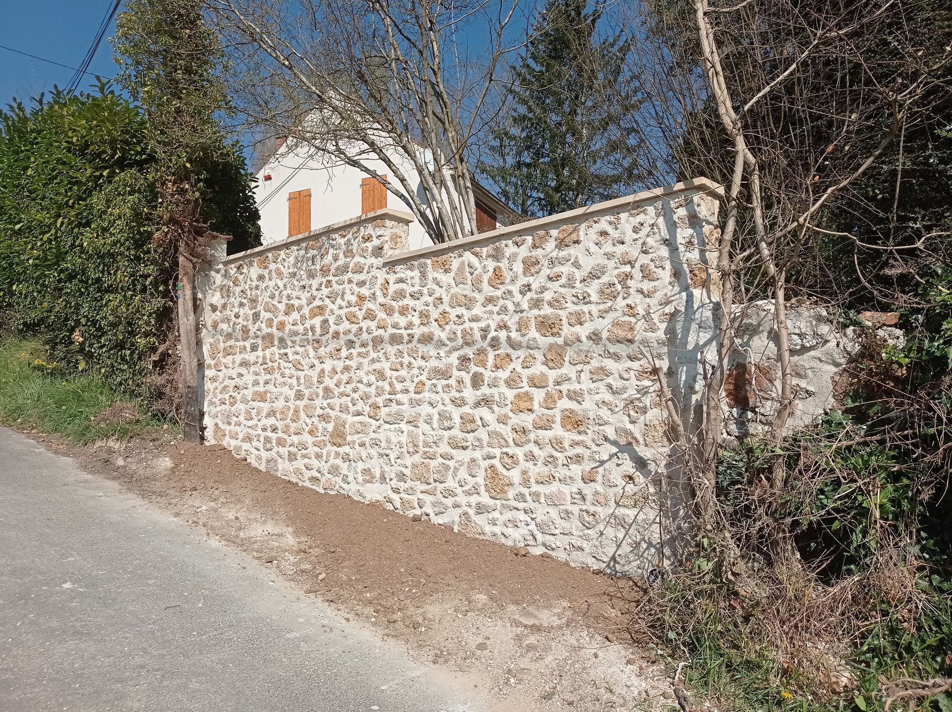 Un mur de pierre claire, avec des arbres et un bâtiment blanc visibles en arrière-plan.