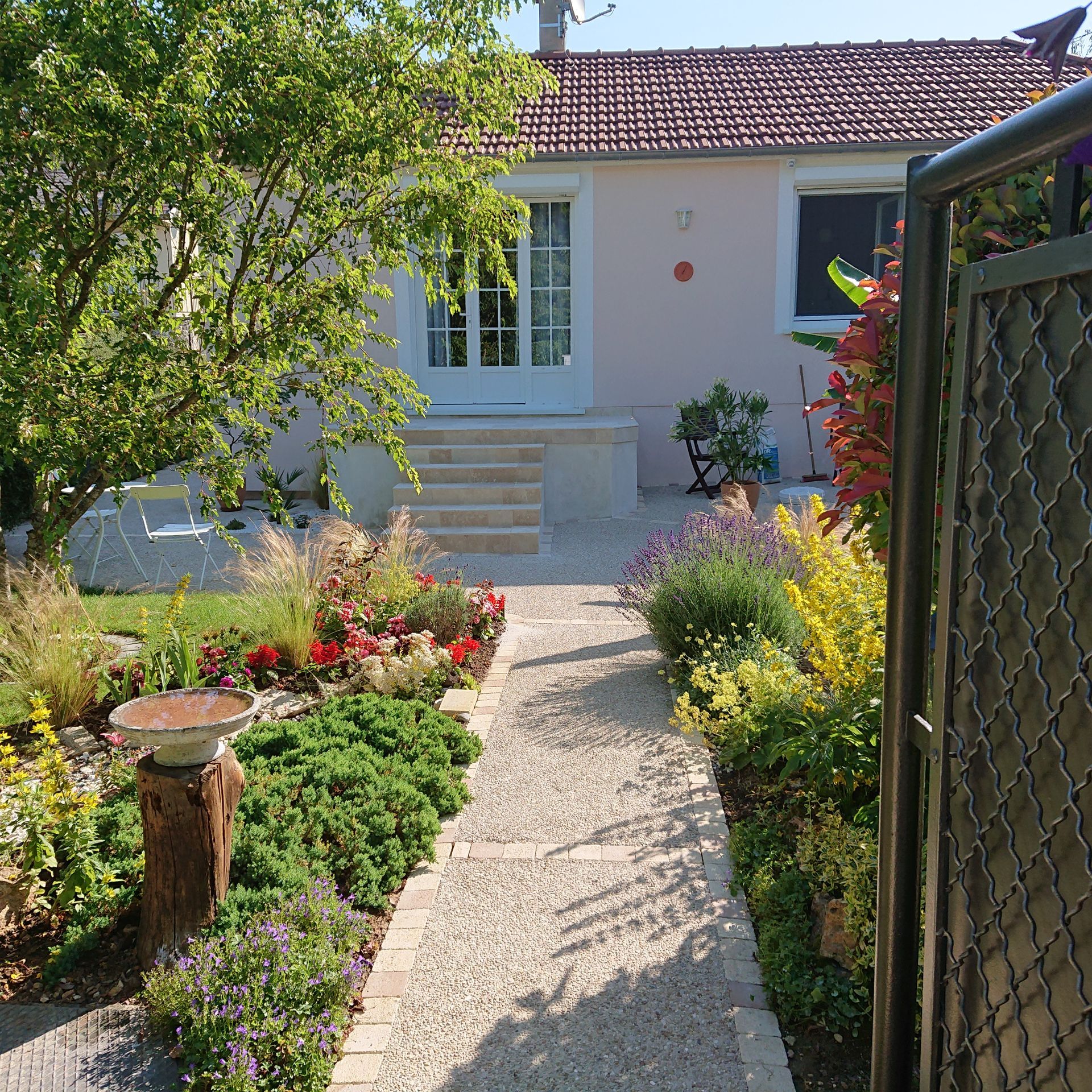 Chemin menant &agrave; une maison rose au toit de tuiles rouges, entour&eacute;e de parterres de jardin color&eacute;s, vue depuis un portail.