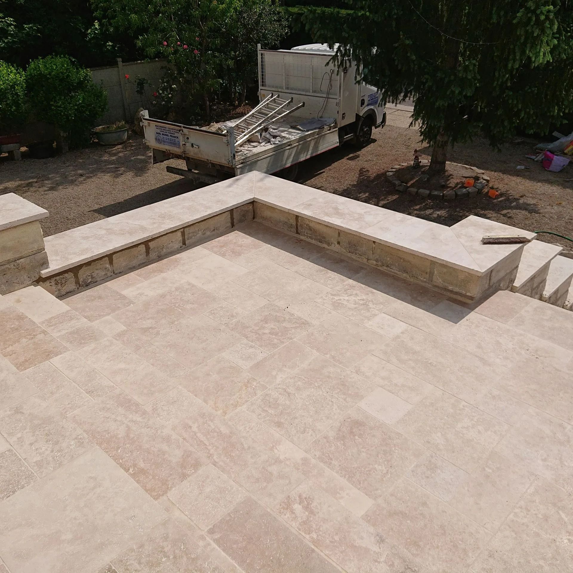 Vue de dessus d'une terrasse en pierre beige avec un muret en forme de L et un camion blanc garé en arrière-plan.