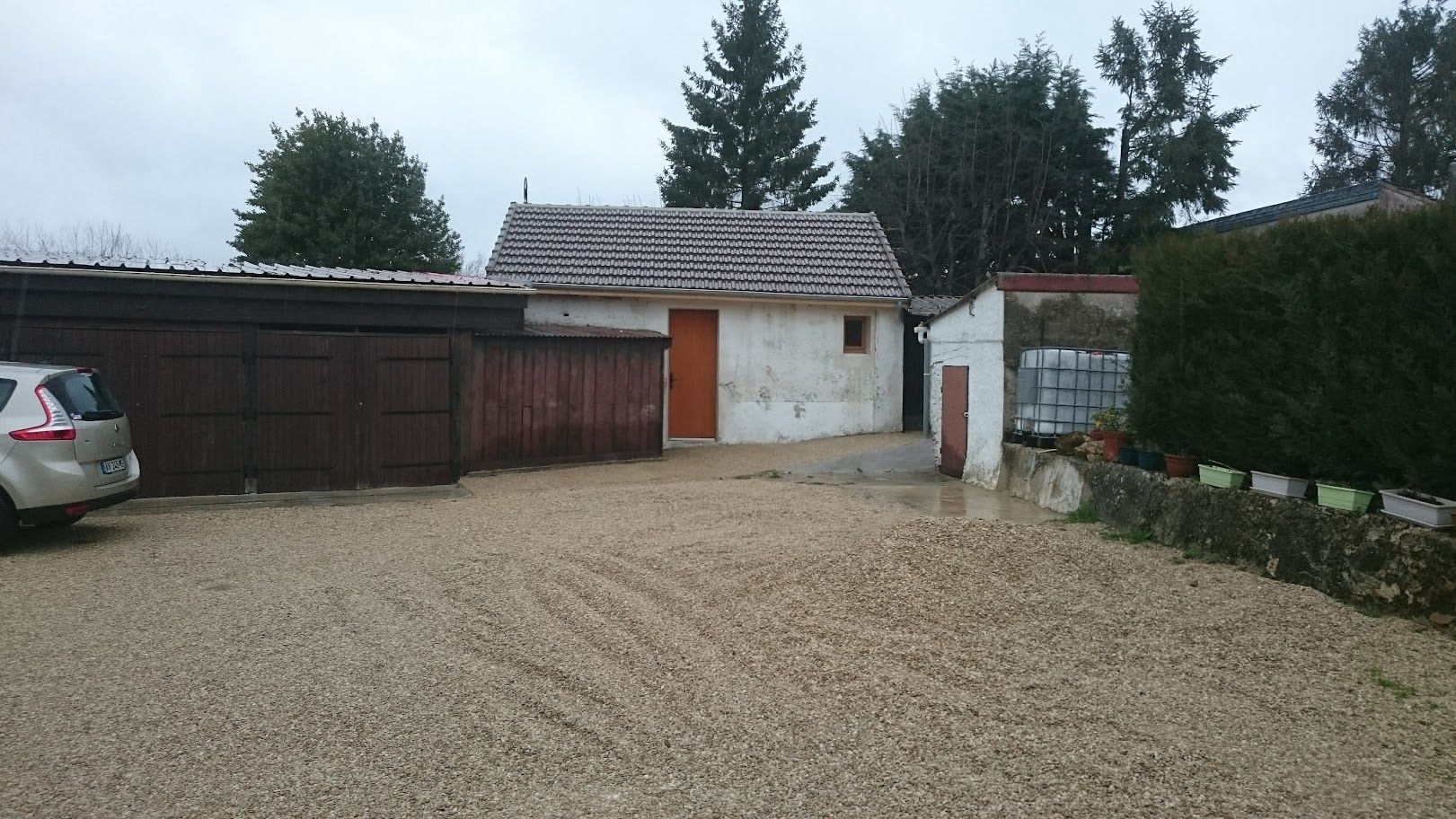 Une allée de gravier mène à une dépendance aux murs blancs délabrés, avec une porte orange.