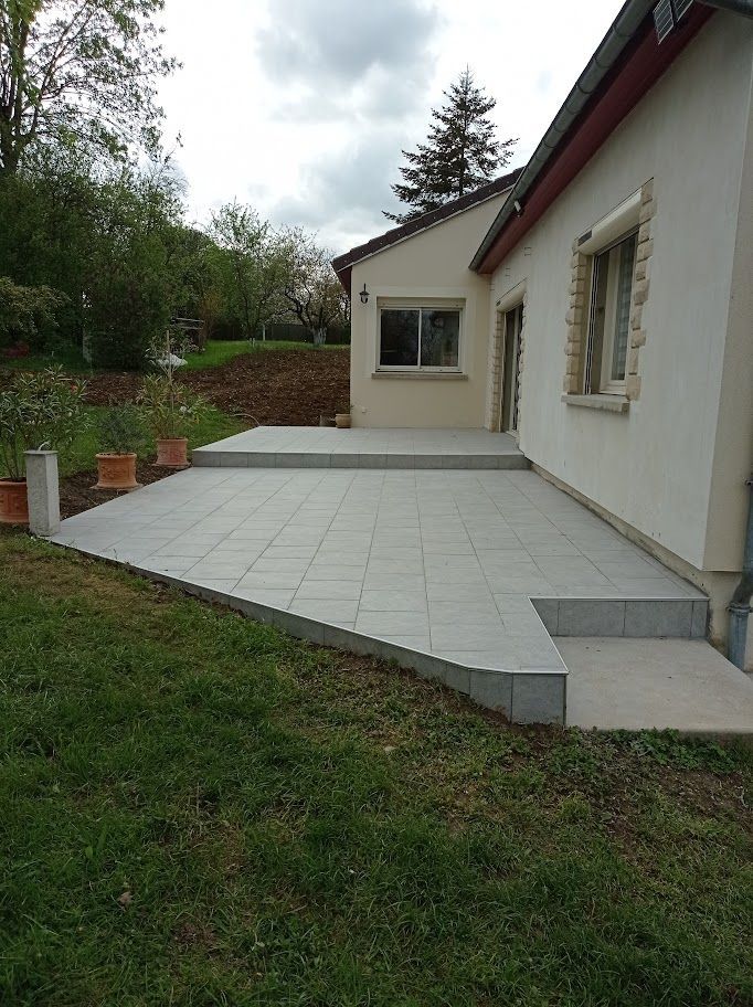 Une terrasse moderne carrelée gris clair avec un dénivelé, attenante à la façade d'une maison de couleur claire.