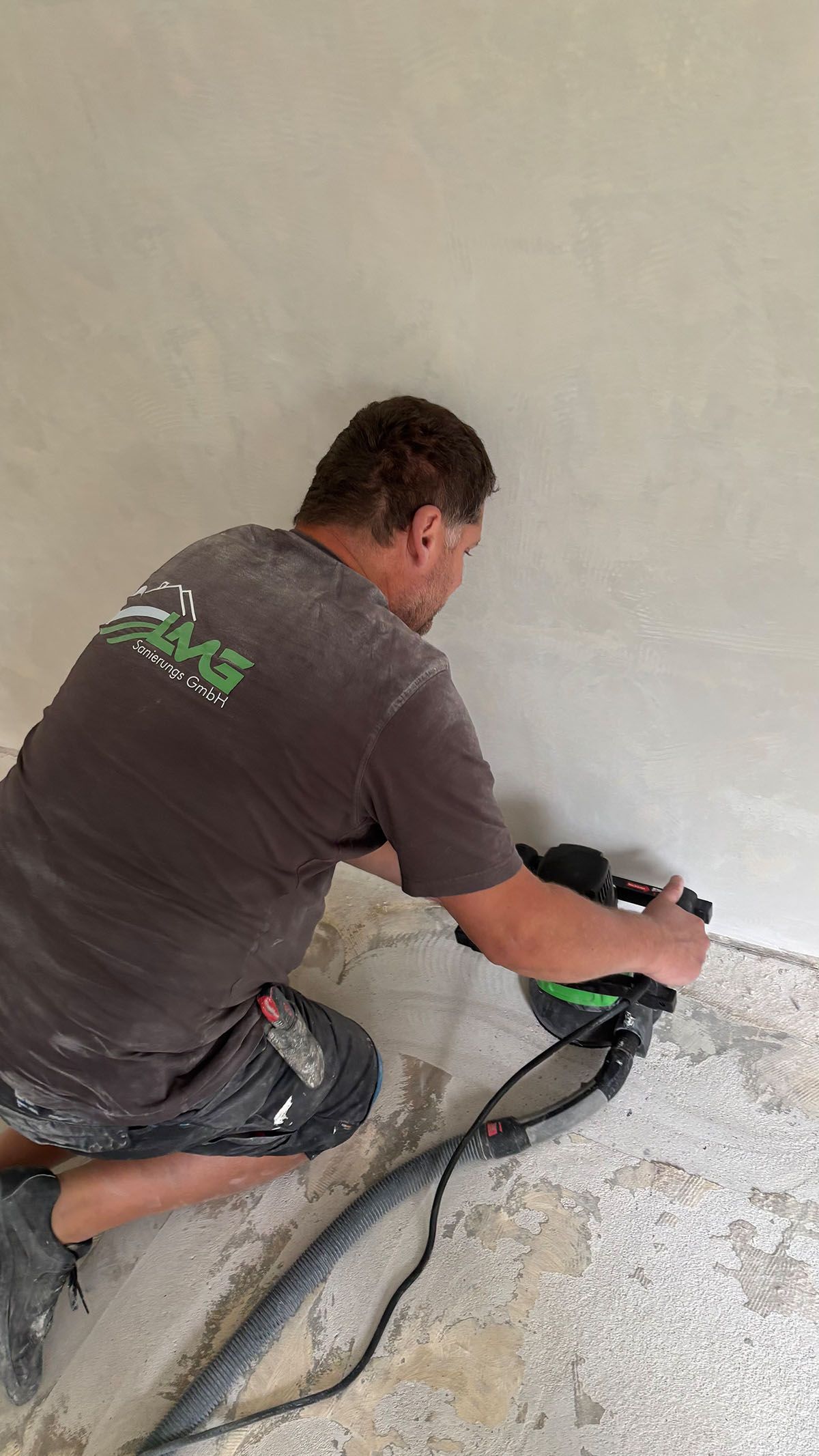 Mann schleift eine Wand, kniet auf einem staubigen Boden und verwendet einen handgeführten elektrischen Schleifer, drinnen.