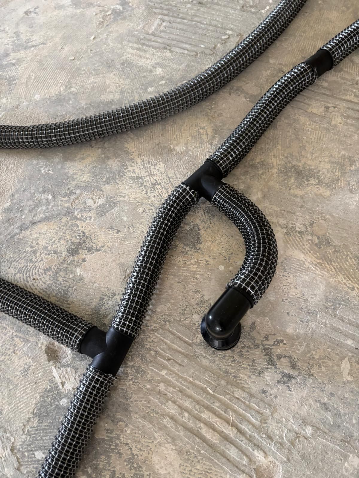 Graue geflochtene Schläuche mit schwarzen Anschlüssen auf einer Betonoberfläche.