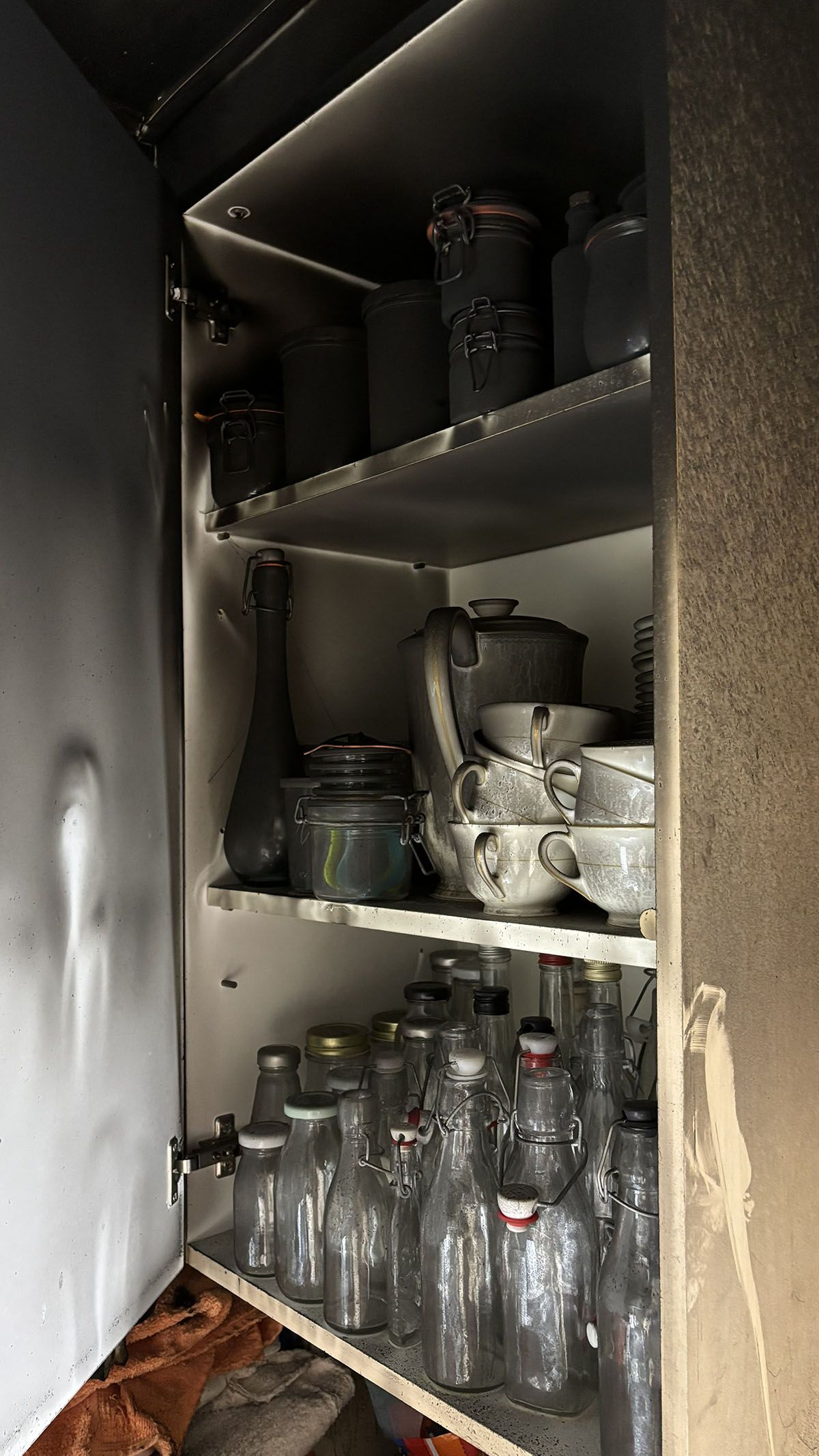 Verbranntes Schrankinneres mit Glasgefäßen, Flaschen und einer rußbedeckten Teekanne.