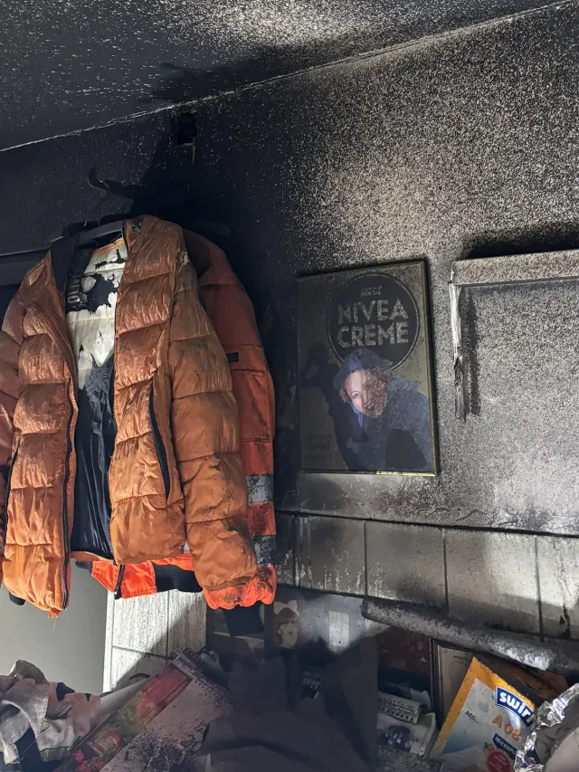Ausgebranntes Zimmerinneres mit Jacken, Nivea-Creme-Aufdruck und Schutt. Rauch und Ruß bedecken die Oberflächen.