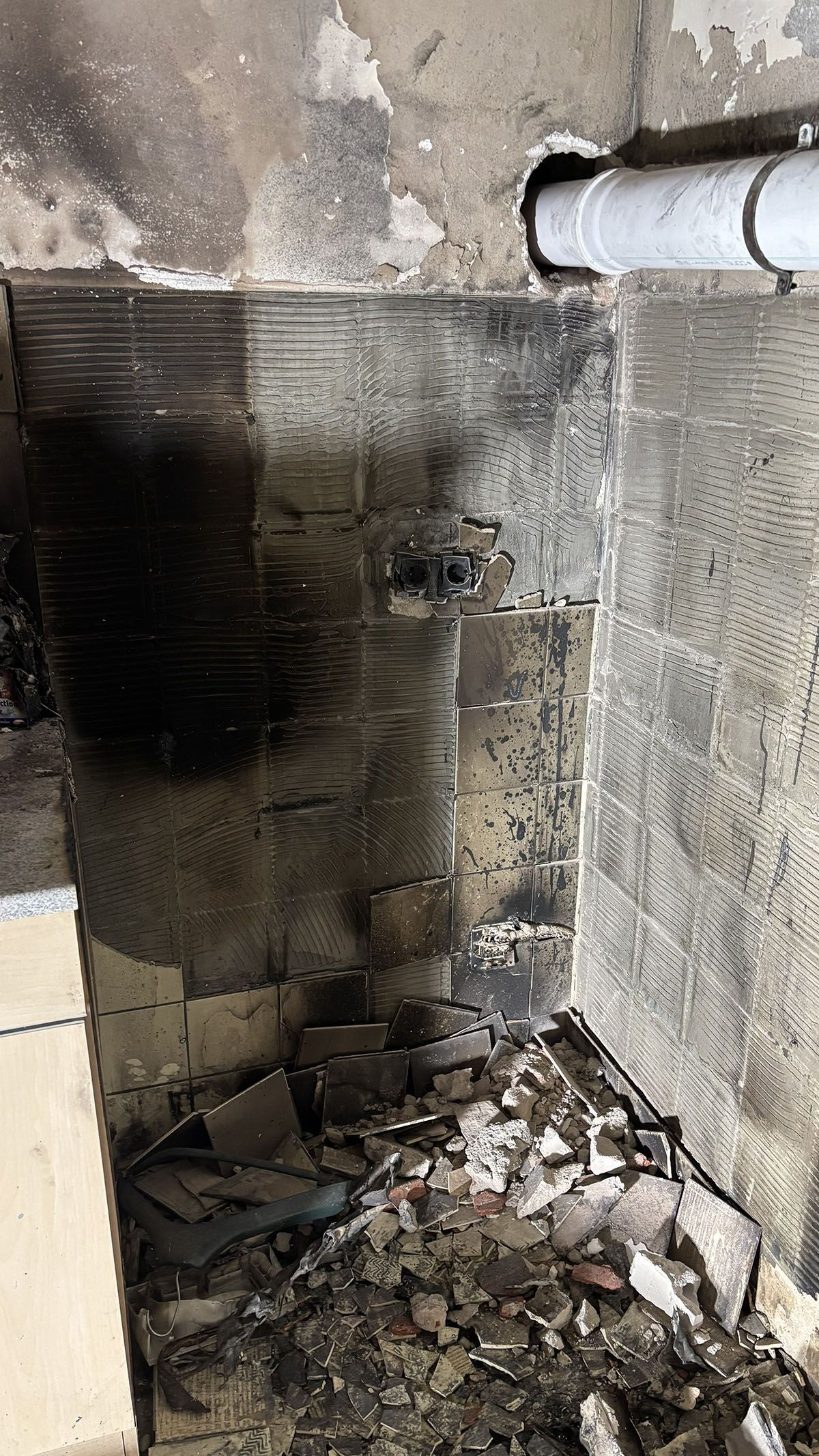 Innenansicht eines beschädigten Bereichs mit verkohlten Wänden und Schutt, vermutlich durch einen Brand. Ein weißes Rohr ist sichtbar.