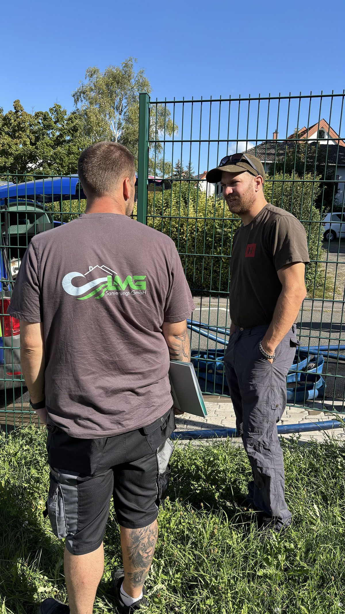 Zwei Männer unterhalten sich an einem sonnigen Tag in der Nähe eines Metallzauns. Einer trägt ein graues Hemd mit einem Logo, der andere einen Hut.