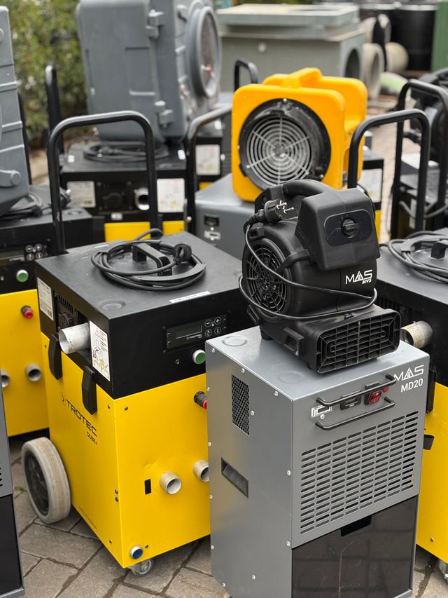 Nahaufnahme einer grauen MAS MD20-Maschine mit einem roten Netzschalter, einer Entlüftung und einem schwarzen Schlauch vor einem Betonboden.