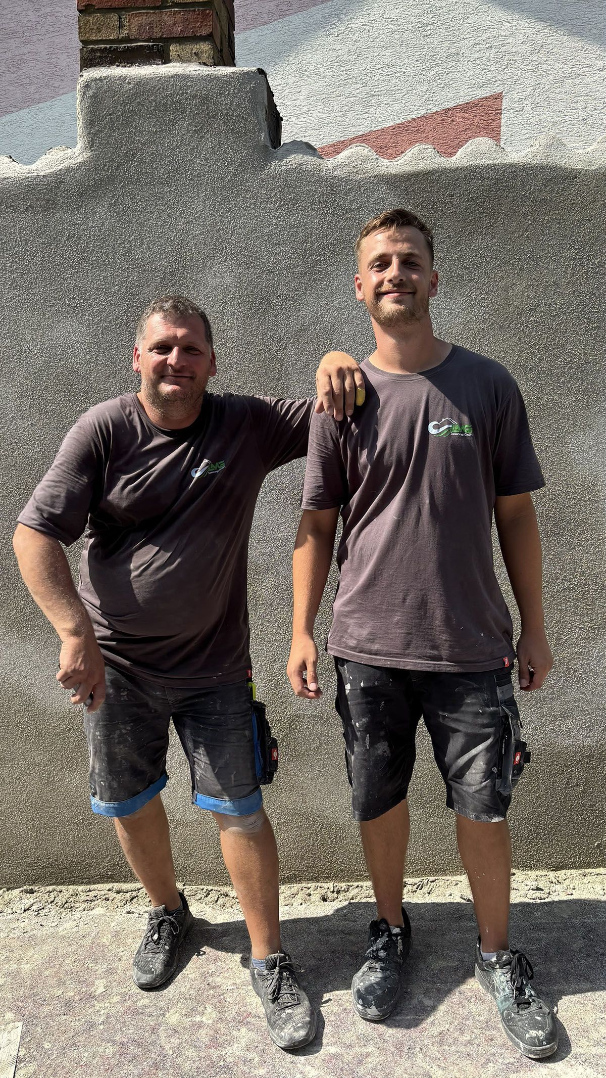 Zwei Männer in Arbeitskleidung posieren vor einer strukturierten Wand, einer hat den Arm auf der Schulter des anderen.