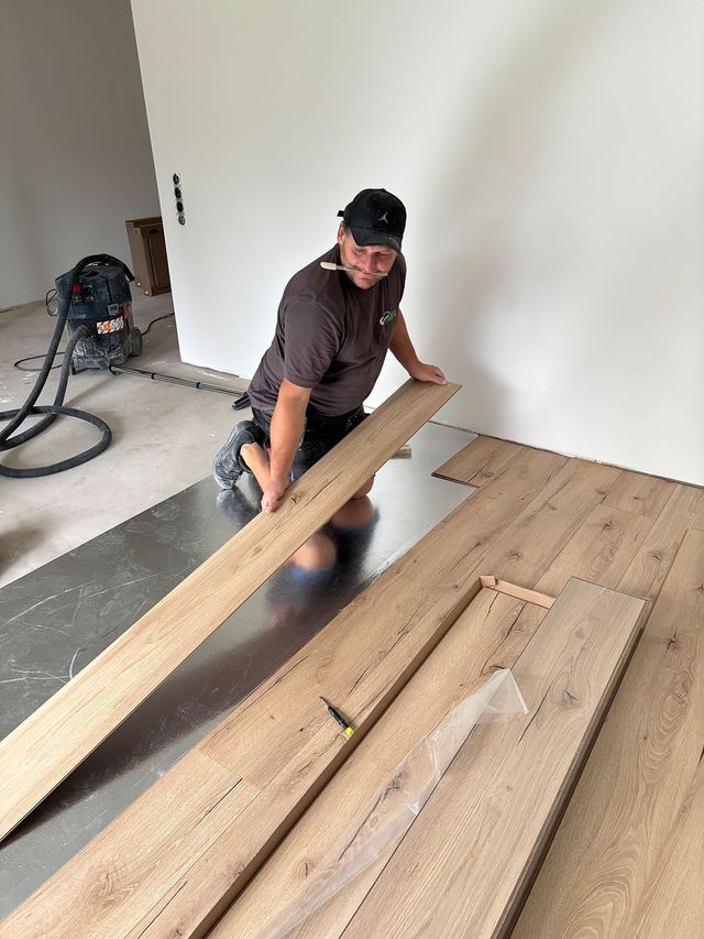 Mann verlegt Holzdielen, kniet im Haus, neben einer weißen Wand und einem Staubsauger.