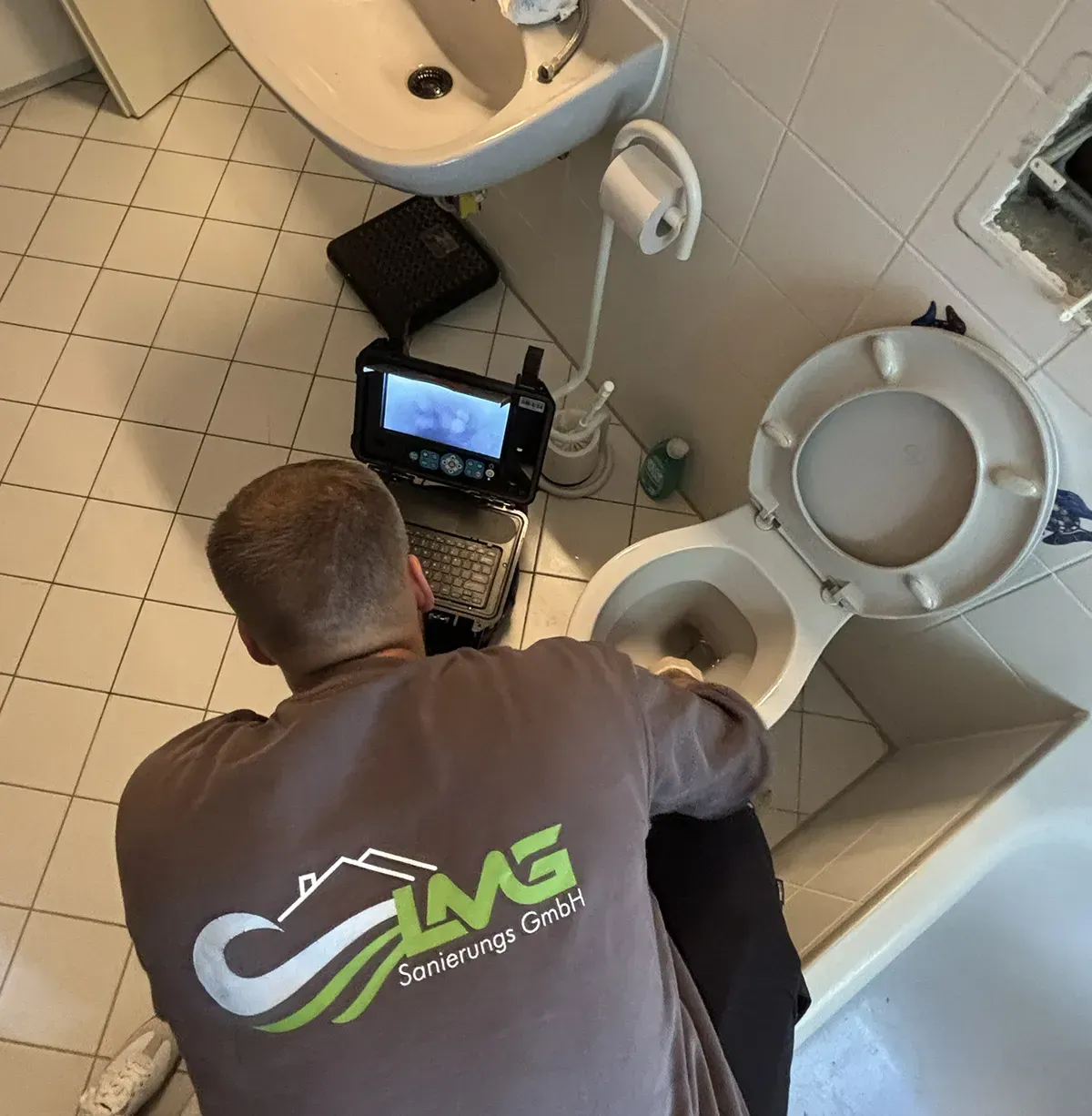 Klempner inspiziert Toilette im Badezimmer mit Kamera. Er trägt ein braunes Hemd mit einem Logo.
