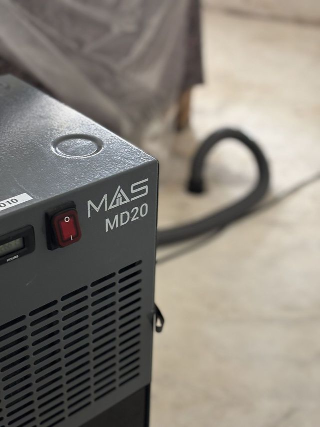 Nahaufnahme einer grauen MAS MD20-Maschine mit einem roten Netzschalter, einer Entlüftung und einem schwarzen Schlauch vor einem Betonboden.