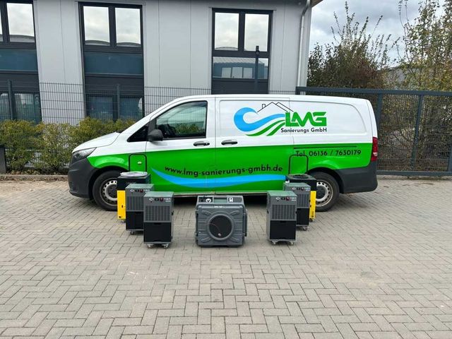 Weißer Lieferwagen mit Logo, geparkt vor einem Gebäude. Vor dem Lieferwagen sind mehrere industrielle Luftentfeuchter aufgestellt.