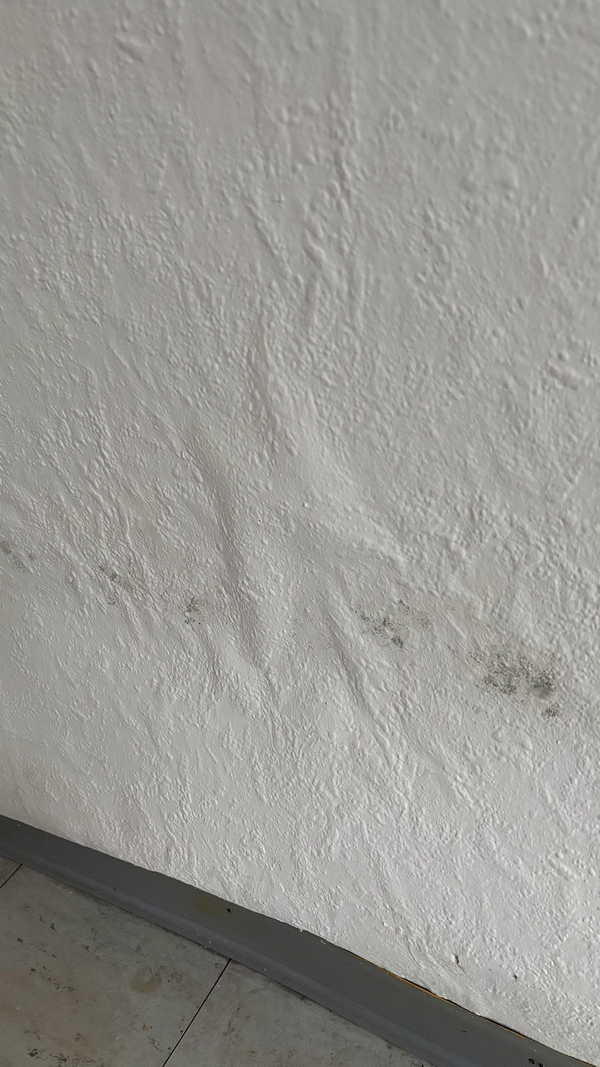 Weiße strukturierte Wand mit dunkleren Flecken, möglicherweise Wasserschäden, in der Nähe einer grauen Zierleiste.